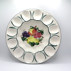 May include: A white ceramic egg plate with 12 individual indentations for serving eggs. The plate features a green border and a colorful fruit design in the center.