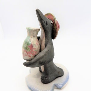 May include: A ceramic penguin figurine wearing a red hat and holding a small, colourful vase. The penguin is standing on a white ceramic base.