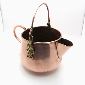 May include: A copper vessel with a rounded body, a side handle, and a tall, arched swing handle attached by brass mounts shaped like human faces. The vessel features a wide, open top and a prominent pouring spout on the right side, showcasing a polished metallic finish.