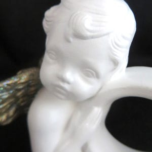 Pu&ograve; includere: Statuetta di cherubino in ceramica bianca con un'espressione serena. Il cherubino ha i capelli ricci e un viso piccolo e dettagliato. La statuetta ha un'ala dorata e iridescente. Lo sfondo &egrave; nero.