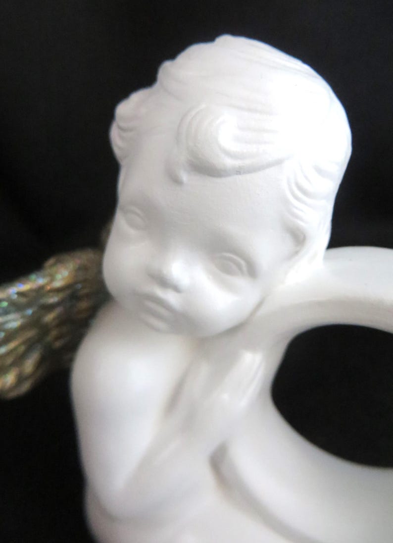 Pu&ograve; includere: Statuetta di cherubino in ceramica bianca con un'espressione serena. Il cherubino ha i capelli ricci e un viso piccolo e dettagliato. La statuetta ha un'ala dorata e iridescente. Lo sfondo &egrave; nero.