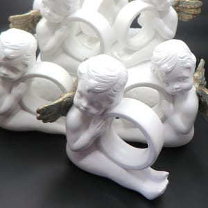 Pu&ograve; includere: Un gruppo di figurine di cherubini bianchi, ognuna con un anello bianco. I cherubini hanno lineamenti dettagliati e piccole ali dorate. Le figurine sono disposte in un gruppo decorativo, adatto per l'arredamento della casa.