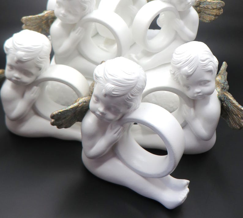 Pu&ograve; includere: Un gruppo di figurine di cherubini bianchi, ognuna con un anello bianco. I cherubini hanno lineamenti dettagliati e piccole ali dorate. Le figurine sono disposte in un gruppo decorativo, adatto per l'arredamento della casa.