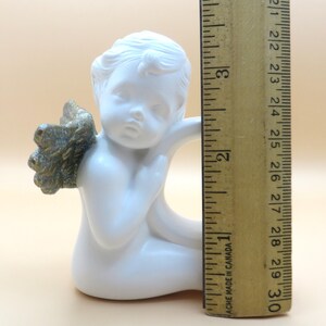 Pu&ograve; includere: Una statuetta di angelo in ceramica bianca con ali color oro. L'angelo &egrave; in posizione seduta, con un'espressione pensosa. La statuetta &egrave; alta circa 7,6 cm.