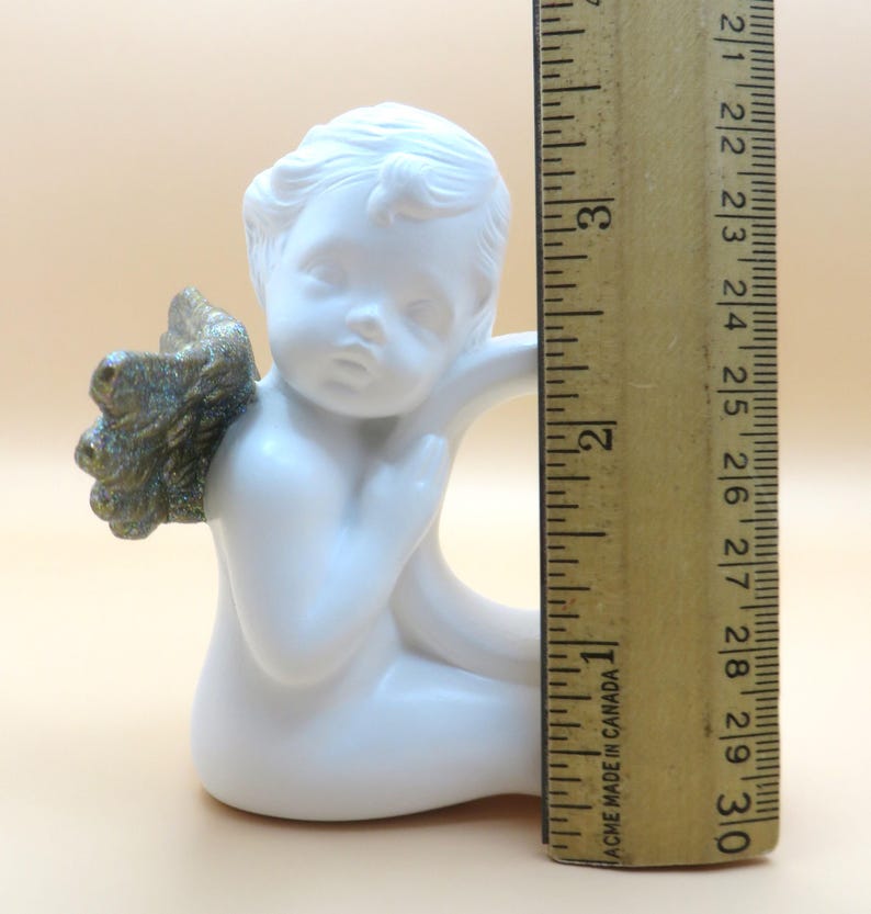 Pu&ograve; includere: Una statuetta di angelo in ceramica bianca con ali color oro. L'angelo &egrave; in posizione seduta, con un'espressione pensosa. La statuetta &egrave; alta circa 7,6 cm.