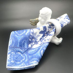 Pu&ograve; includere: Un portatovagliolo a cherubino in ceramica bianca con ali color oro, che tiene un tovagliolo a fantasia blu e bianco. Il tovagliolo presenta un disegno floreale e paisley. Il cherubino &egrave; su una superficie scura.