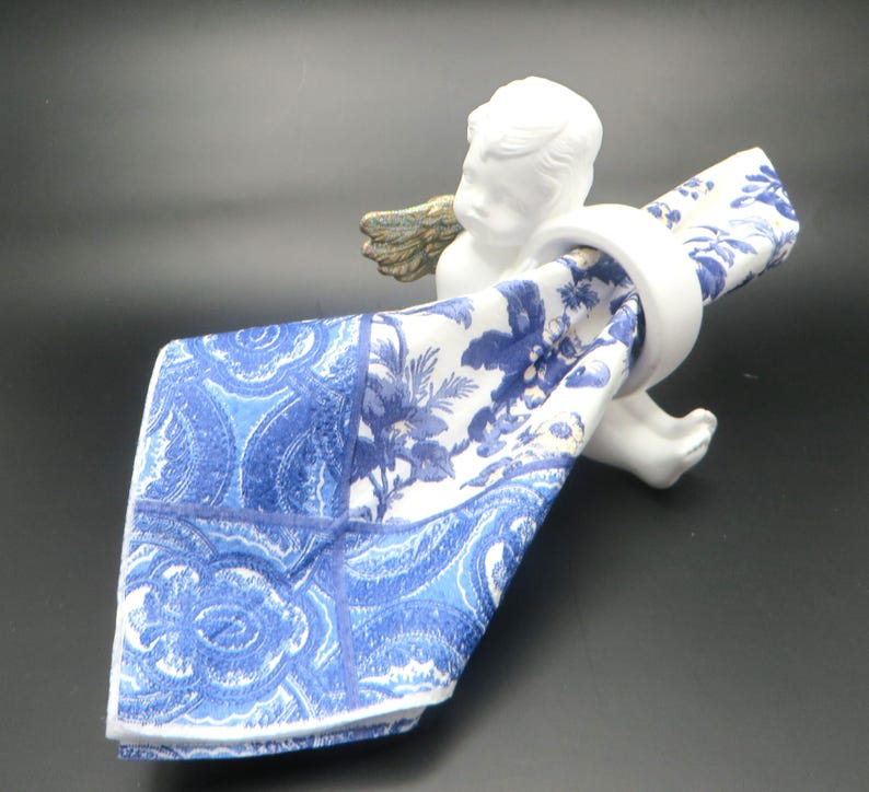 Pu&ograve; includere: Un portatovagliolo a cherubino in ceramica bianca con ali color oro, che tiene un tovagliolo a fantasia blu e bianco. Il tovagliolo presenta un disegno floreale e paisley. Il cherubino &egrave; su una superficie scura.