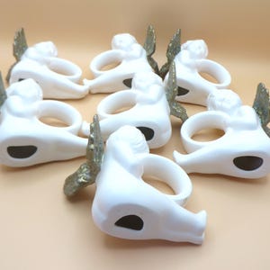 Pu&ograve; includere: Una collezione di sette portatovaglioli a forma di angelo in ceramica bianca. Ogni anello presenta un angelo reclinato con ali color oro. I portatovaglioli hanno un'apertura circolare per un tovagliolo e un'apertura ovale scura sul fondo.