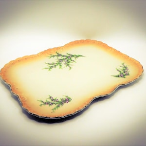 May include: A vintage serving tray in off-white with a scalloped edge and an orange border. It features sprigs of purple and green flowers. A decorative piece for serving or display, with a classic design.