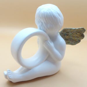 Pu&ograve; includere: Una statuetta di cherubino bianco che tiene un anello bianco. Il cherubino ha ali color oro ed &egrave; seduto con la schiena rivolta allo spettatore. La statuetta &egrave; realizzata in un materiale liscio e opaco.