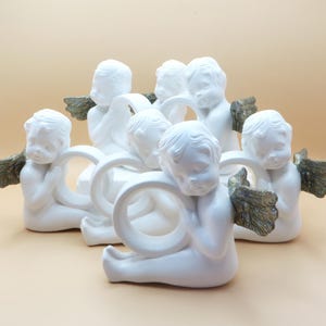 Pu&ograve; includere: Una collezione di figurine di cherubini bianchi, ognuna con un anello e ali color oro. I cherubini sono disposti in un motivo circolare, creando un'esposizione decorativa.