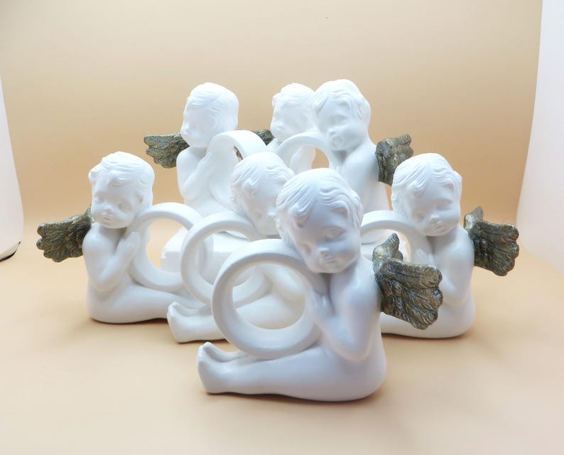 Pu&ograve; includere: Una collezione di figurine di cherubini bianchi, ognuna con un anello e ali color oro. I cherubini sono disposti in un motivo circolare, creando un'esposizione decorativa.