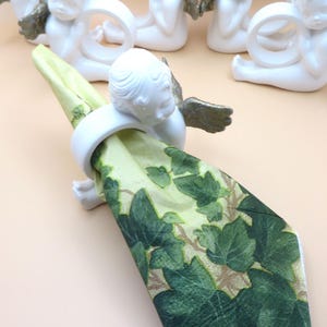 Pu&ograve; includere: Portatovaglioli a forma di cherubini bianchi con ali color oro che tengono un tovagliolo verde e beige con un motivo a foglie. I cherubini sono seduti. I portatovaglioli sono decorativi e adatti a una tavola formale.