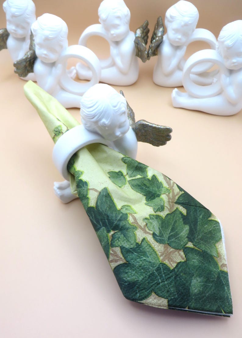 Pu&ograve; includere: Portatovaglioli a forma di cherubini bianchi con ali color oro che tengono un tovagliolo verde e beige con un motivo a foglie. I cherubini sono seduti. I portatovaglioli sono decorativi e adatti a una tavola formale.