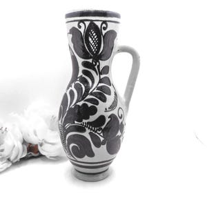 May include: A white ceramic pitcher with a black floral design and a handle. The pitcher has a narrow neck and a wider base. The floral design includes stylized leaves and flowers. The pitcher is likely handmade and has a rustic, folk art aesthetic.
