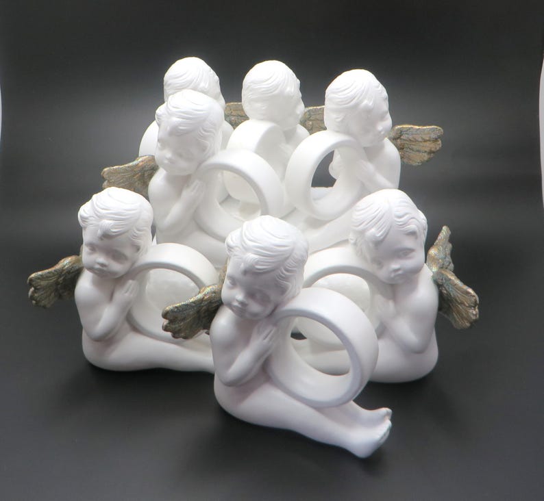 Pu&ograve; includere: Un centrotavola in ceramica bianca con otto figure di cherubini. Ogni cherubino tiene un anello bianco, con ali color oro. I cherubini sono disposti in un motivo circolare, creando un design decorativo ed elegante.