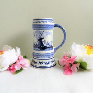 May include: A blue and white ceramic beer stein featuring a Dutch windmill scene. The stein has a wide base and a handle.