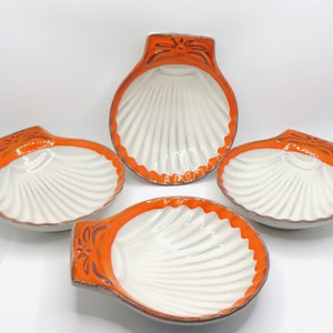 May include: A set of four ceramic dishes shaped like scallop shells. Each dish features a glossy white interior with embossed ridges and a vibrant orange border along the outer edge, accented with dark brown decorative line details at the top.