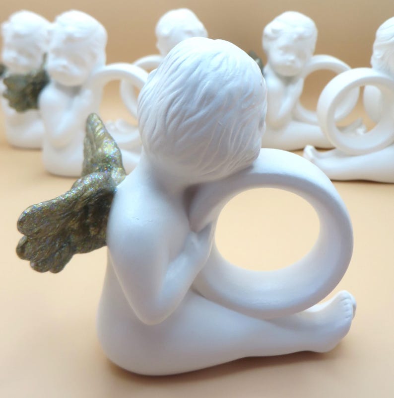 Pu&ograve; includere: Portatovaglioli a forma di angelo in ceramica bianca con ali dorate. Gli angeli sono seduti e tengono un anello. Le ali hanno una finitura strutturata e metallica. Sono visibili diversi anelli, che suggeriscono un set.