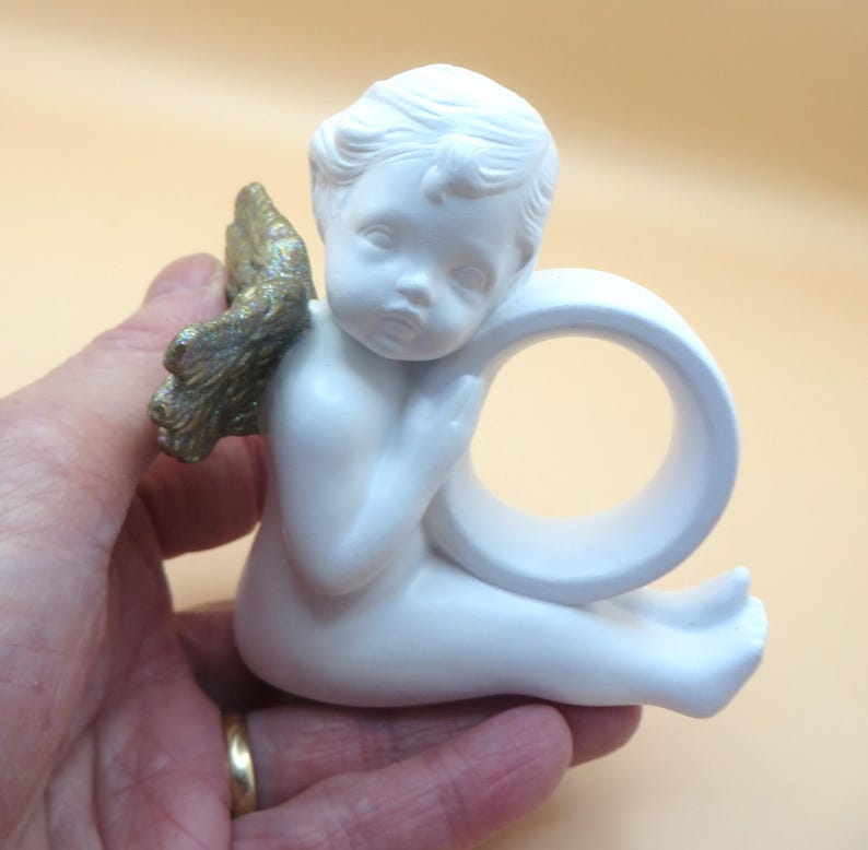 Pu&ograve; includere: Statuina di angelo in ceramica bianca con ali color oro. L'angelo &egrave; in posizione seduta, con un design a forma di anello incorporato. La statuina &egrave; tenuta in mano.