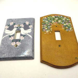 May include: Two decorative light switch covers. One is grey with a dove and floral design. The other is wooden with a floral design. Both have a single switch opening and screw holes for mounting.