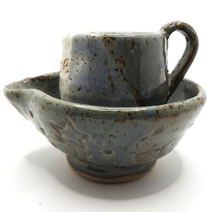 May include: A handmade ceramic mug and bowl set featuring a speckled blue-grey glaze with brown accents. The bowl has a wide rim with a pouring spout and embossed star patterns, while the mug has a sturdy handle and a partial white glazed section.