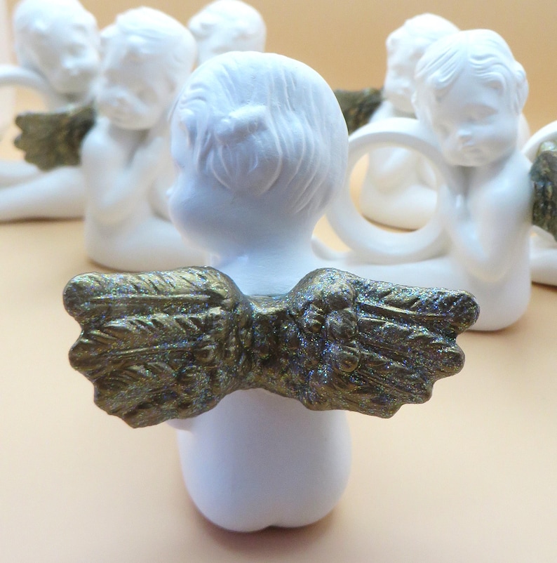 Pu&ograve; includere: Statuine di angeli in ceramica bianca con ali color oro. Gli angeli sono in posizione seduta, con le ali spiegate. Le ali hanno una finitura strutturata e glitterata. Sono visibili diverse statuine, che suggeriscono un set o una collezione.