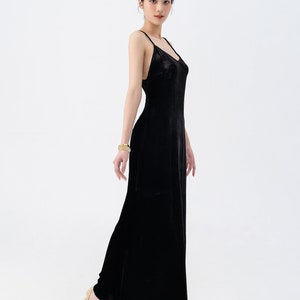 May include: A woman wearing a black velvet maxi dress with spaghetti straps. She is standing on a white background and wearing gold bracelets and nude heels.