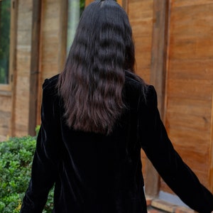 Pode incluir: Uma mulher vestindo um blazer de veludo preto de mangas compridas. Ela est&aacute; em p&eacute; em frente a uma porta de madeira.