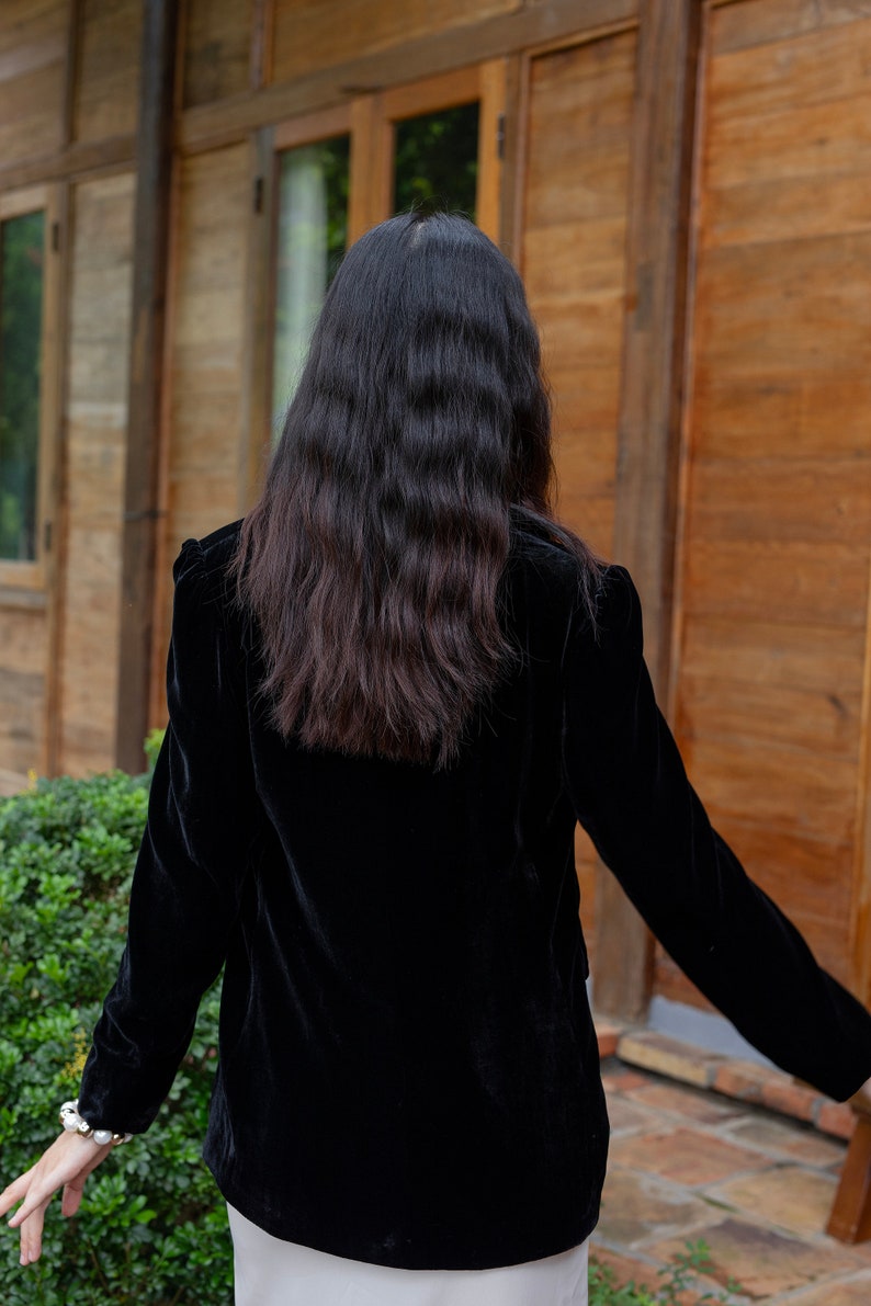 Pode incluir: Uma mulher vestindo um blazer de veludo preto de mangas compridas. Ela est&aacute; em p&eacute; em frente a uma porta de madeira.