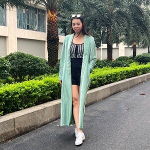 May include: A woman wearing a long, light green velvet cardigan over a black and white checkered top and black shorts. She is wearing white sneakers and has her hair pulled back.