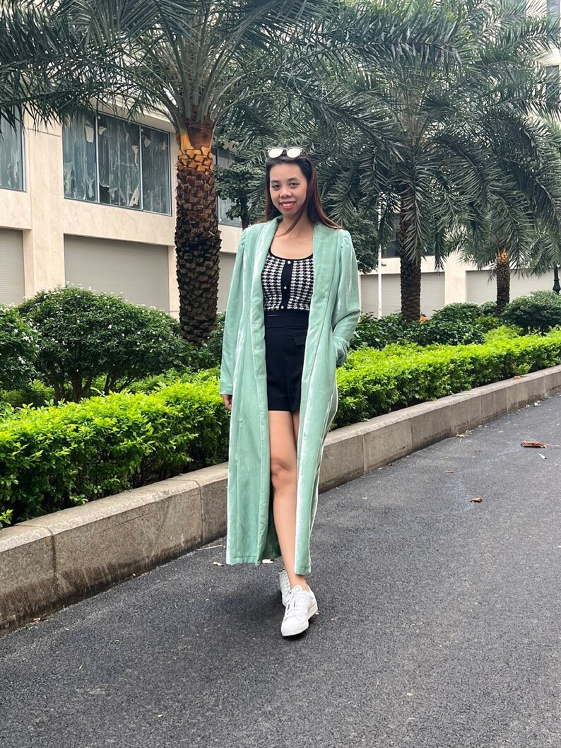 May include: A woman wearing a long, light green velvet cardigan over a black and white checkered top and black shorts. She is wearing white sneakers and has her hair pulled back.