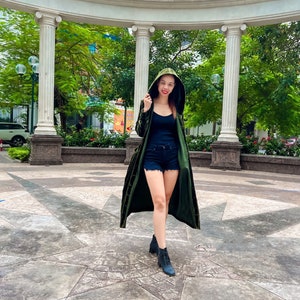 May include: A woman wearing a green velvet hooded jacket with a black tank top and denim shorts. She is standing in a circular courtyard with white columns and green trees.