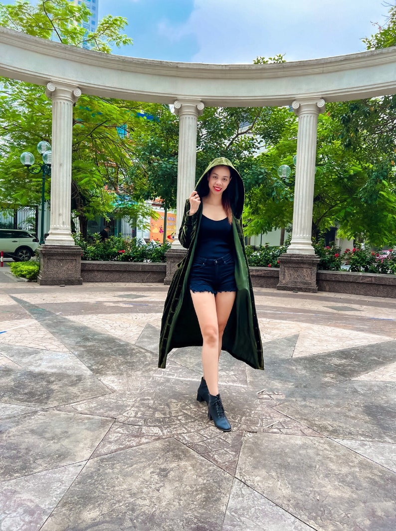 May include: A woman wearing a green velvet hooded jacket with a black tank top and denim shorts. She is standing in a circular courtyard with white columns and green trees.