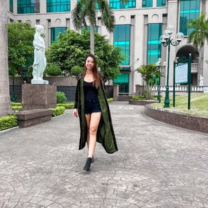 May include: A woman wearing a long, green, velvet jacket with a black tank top and denim shorts. She is walking on a paved path in front of a building with large windows.