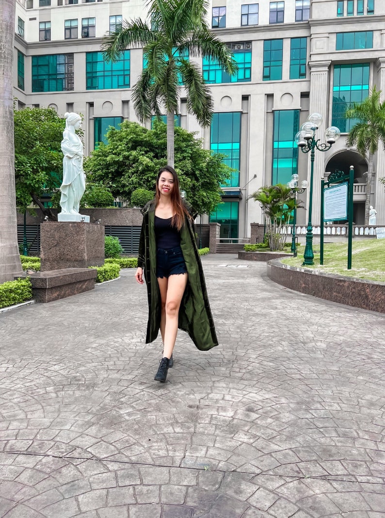 May include: A woman wearing a long, green, velvet jacket with a black tank top and denim shorts. She is walking on a paved path in front of a building with large windows.