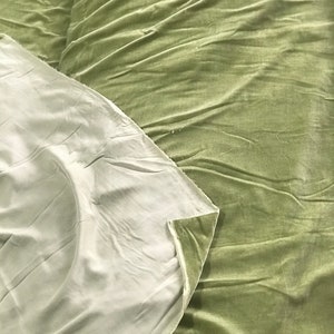 Puede incluir: Primer plano de una tela de terciopelo verde salvia con una tela blanca debajo. La tela est&aacute; doblada y tiene una textura suave y lisa.