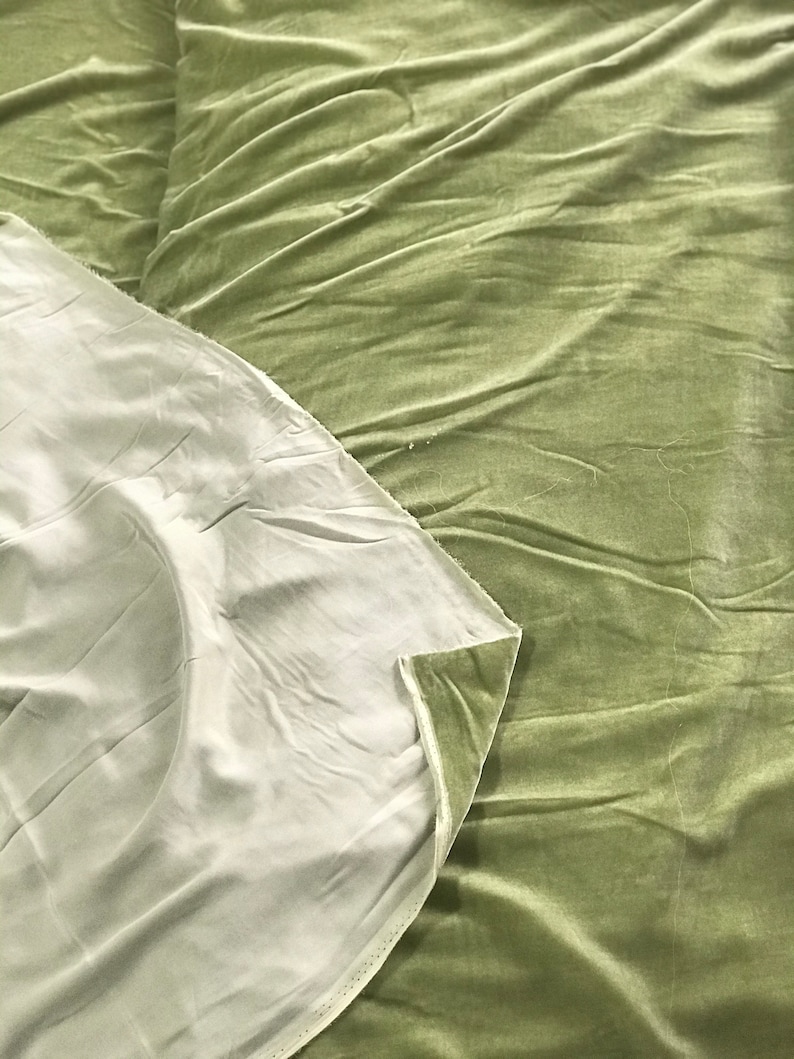 Puede incluir: Primer plano de una tela de terciopelo verde salvia con una tela blanca debajo. La tela est&aacute; doblada y tiene una textura suave y lisa.