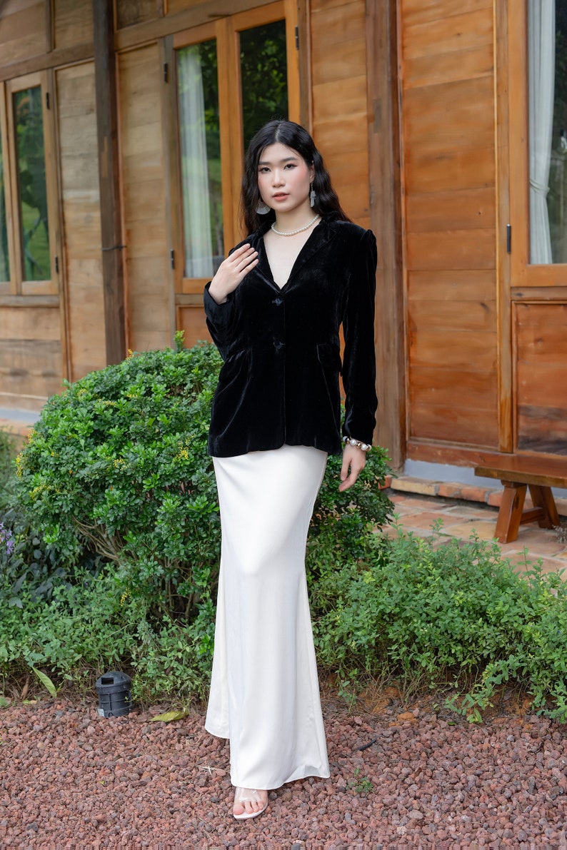 Pode incluir: Uma mulher vestindo um blazer de veludo preto sobre uma saia de cetim branco. Ela est&aacute; em p&eacute; na frente de um pr&eacute;dio de madeira com um arbusto verde atr&aacute;s dela.