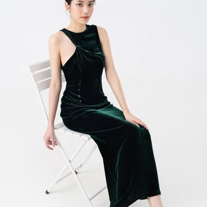 May include: A dark green velvet dress with a unique asymmetrical neckline. The sleeveless dress has a fitted bodice and a floor-length skirt. The model is seated on a white folding chair, wearing nude-colored heels with clear panels.