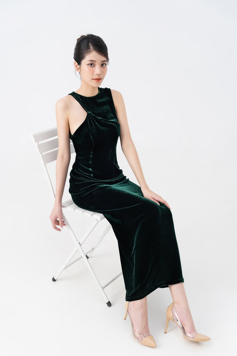May include: A dark green velvet dress with a unique asymmetrical neckline. The sleeveless dress has a fitted bodice and a floor-length skirt. The model is seated on a white folding chair, wearing nude-colored heels with clear panels.