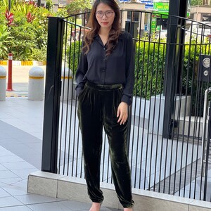May include: A woman wearing a black collared shirt and dark green velvet pants. She is standing in front of a black metal gate.