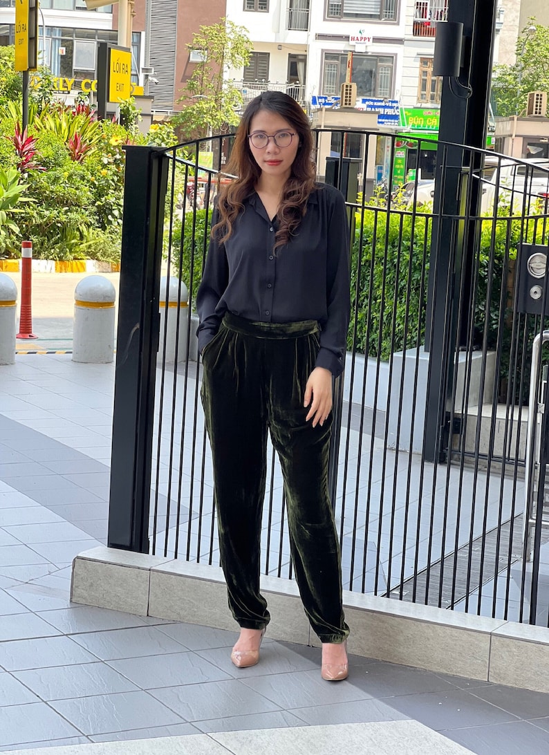 May include: A woman wearing a black collared shirt and dark green velvet pants. She is standing in front of a black metal gate.