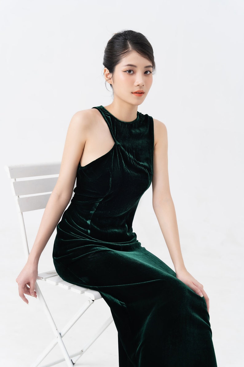 May include: A dark green velvet dress with a unique asymmetrical neckline. The sleeveless gown has a fitted bodice and a floor-length skirt. The model is seated on a white chair against a white background. The dress is a rich, jewel-toned color.