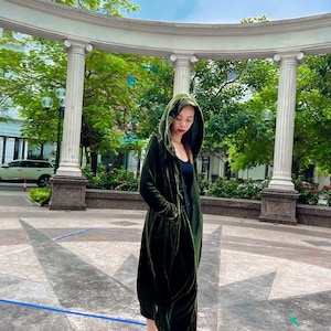 May include: A woman wearing a long, dark green velvet hooded coat with a cinched waist and pockets. She is standing in a courtyard with a white archway and green trees in the background.