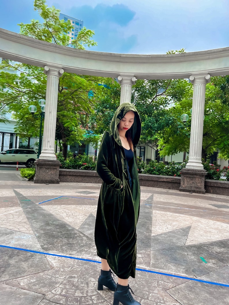 May include: A woman wearing a long, dark green velvet hooded coat with a cinched waist and pockets. She is standing in a courtyard with a white archway and green trees in the background.
