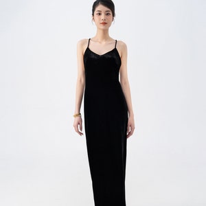 May include: A woman wearing a black velvet maxi dress with thin straps. She is standing on a white background and wearing nude heels.
