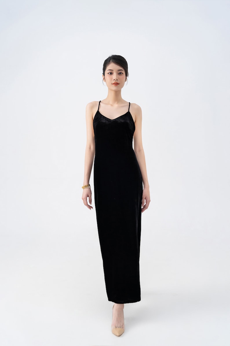 May include: A woman wearing a black velvet maxi dress with thin straps. She is standing on a white background and wearing nude heels.