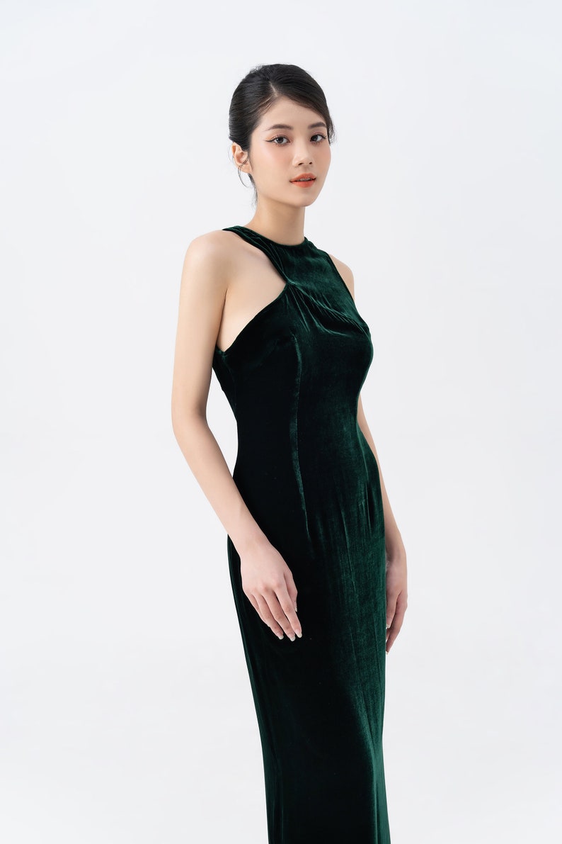 May include: A dark green velvet gown with a high neckline and a unique shoulder cutout. The dress is form-fitting and floor-length, showcasing a sleek and elegant silhouette. The fabric has a rich, luxurious sheen, perfect for formal events.
