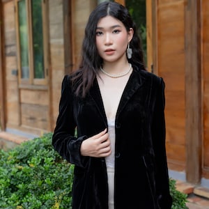 Pode incluir: Uma mulher vestindo um blazer de veludo preto com um colar e brincos de p&eacute;rolas. Ela est&aacute; em p&eacute; na frente de um pr&eacute;dio de madeira com uma saia branca.